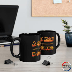 Lifestyle image of A black ceramic coffee mug featuring bold retro orange‑and‑yellow gradient typography that reads “A Hard Man Is Good to Find,” with a vintage horizontal accent line and small star details.